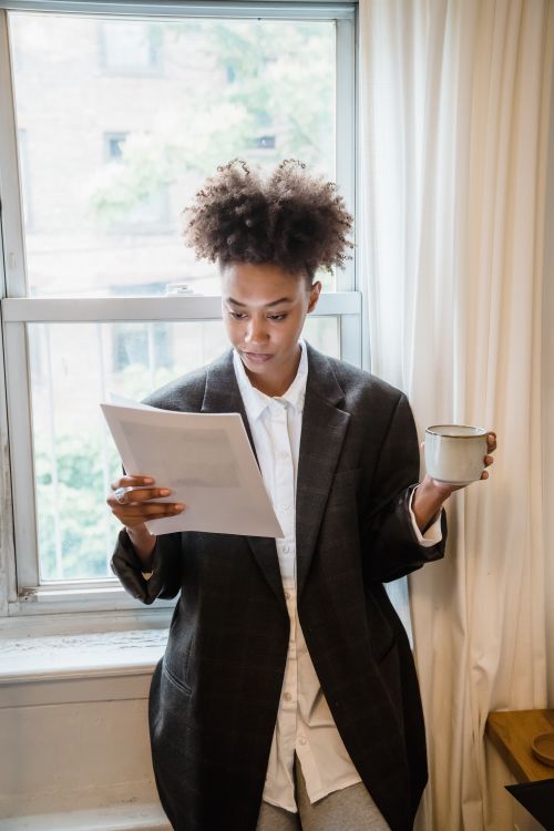 Free A focused woman reads a document while holding a drink by a sunlit window indoors. Stock Photo