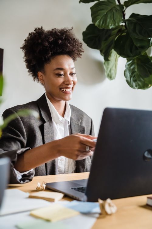 Free Woman with curly hair smiling while working on a laptop at a home office desk. Stock Photo
