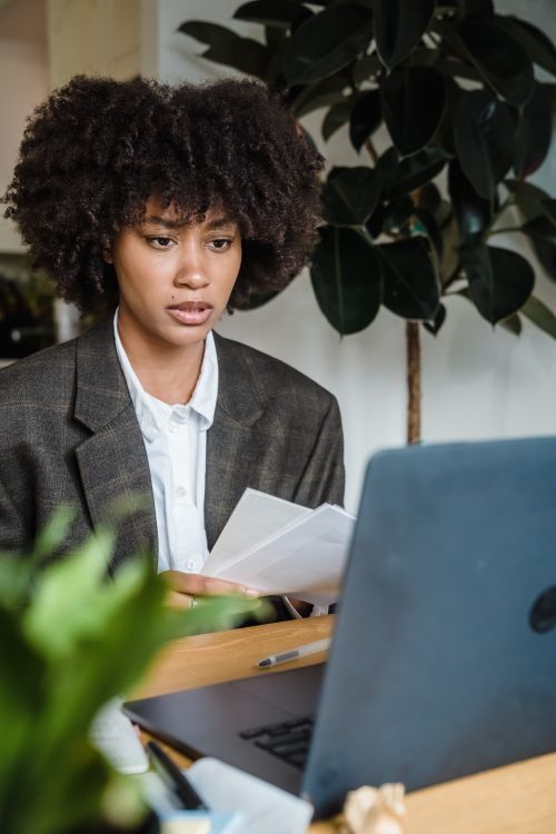 Free Focused businesswoman with curly hair analyzing work documents on laptop, indoors. Stock Photo