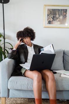 An adult woman looks frustrated while remote working on a sofa with a laptop, documents spread around.