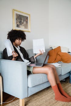 A woman on a sofa managing finances with a laptop and documents, appearing stressed.