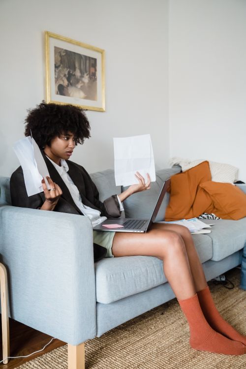 Free A woman on a sofa managing finances with a laptop and documents, appearing stressed. Stock Photo