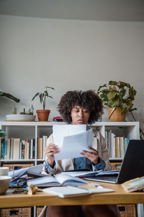 Free A woman reads documents intensely at her home office desk filled with books and a laptop, surrounded by plants. Stock Photo