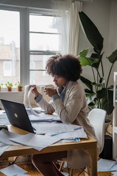 Woman with curly hair working at home office with laptop and documents, sipping coffee.