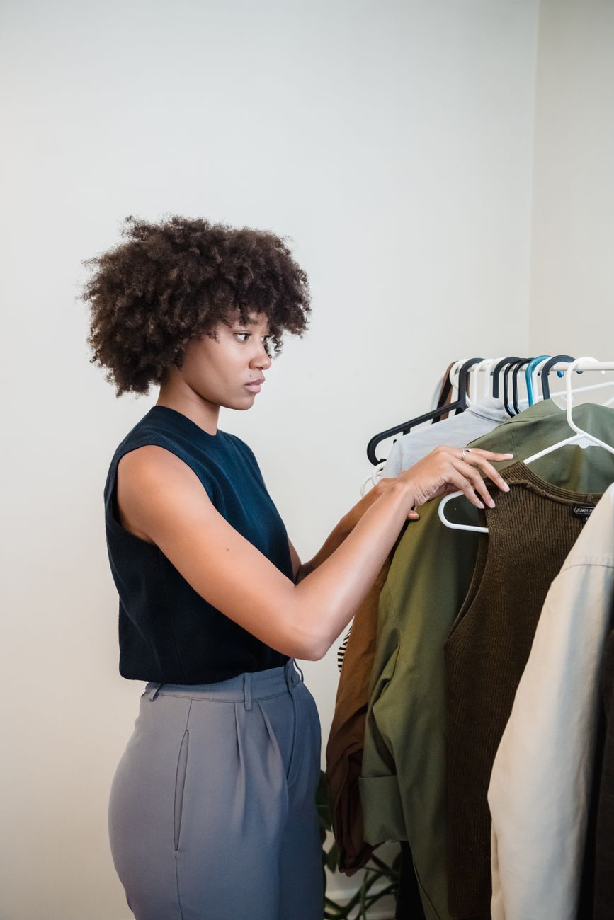 A woman with curly hair selects clothes from a hanger rack in a minimalist indoor setting.