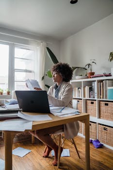 A woman reviewing documents at a home office desk, surrounded by plants and natural light.