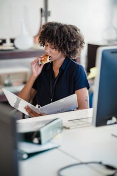 Young woman multitasking by eating a snack and reading documents at her desk.