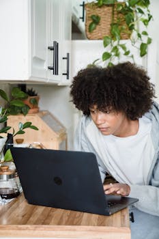 A young woman with curly hair using a laptop in a kitchen surrounded by house plants, creating a homey atmosphere.