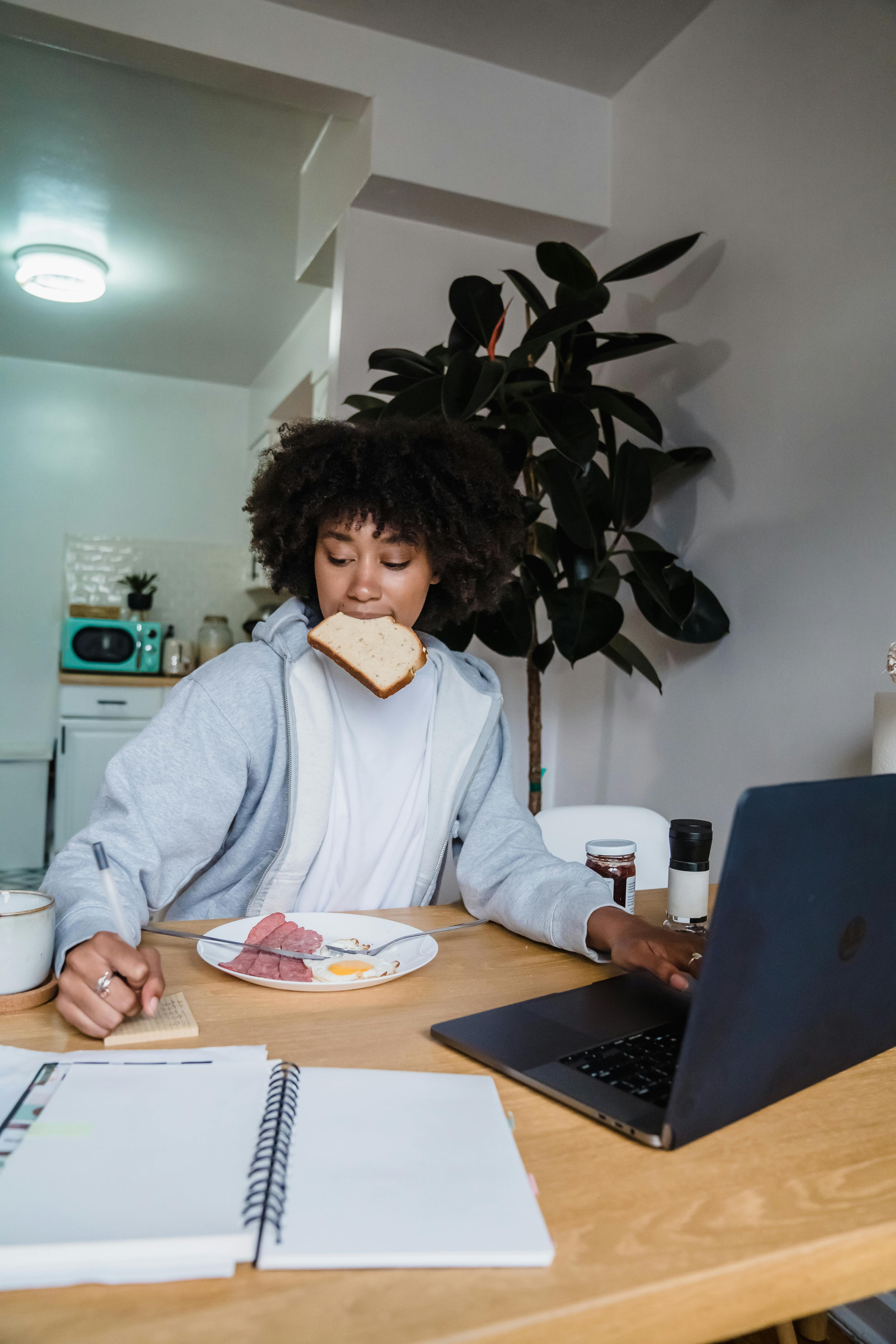 woman eating breakfast writing in a notebook and using laptop at the same time