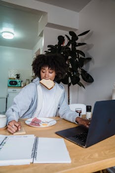 Woman eating breakfast while multitasking with a laptop and notebook indoors.