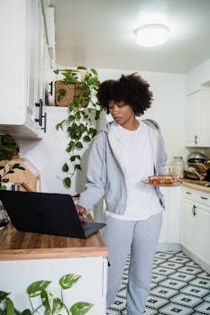 A woman uses her laptop while enjoying breakfast in a cozy home kitchen setting.