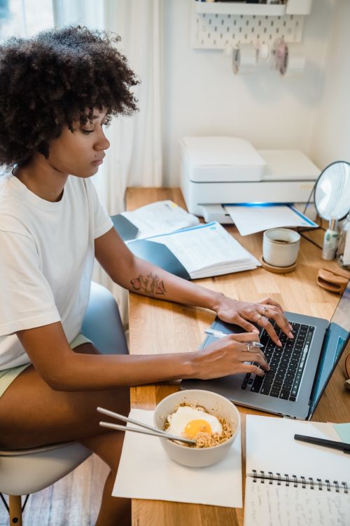 Free Woman using laptop while having breakfast at home, surrounded by books and a notebook. Stock Photo