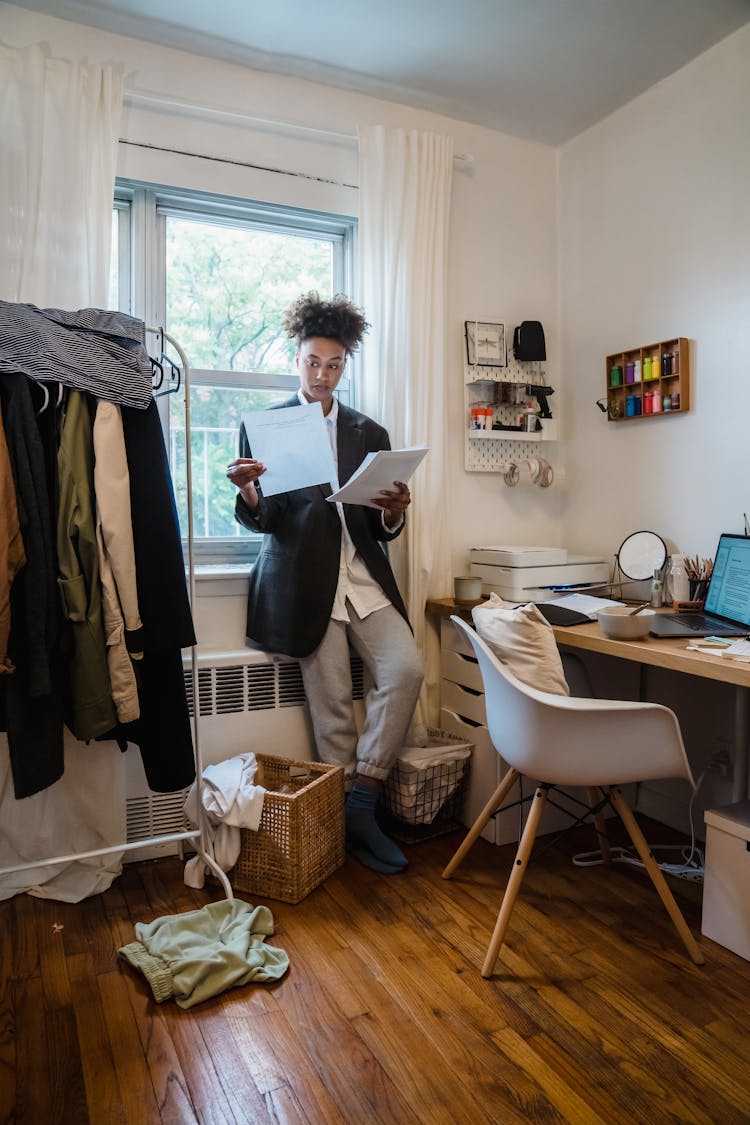 Woman In A Messy Room Standing And Reading Documents With An Open Laptop On The Desk 