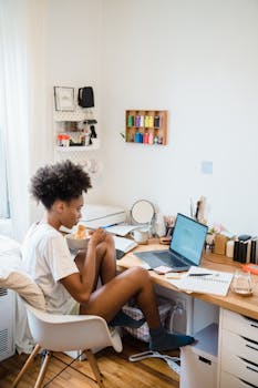 Young woman multitasking at home desk with laptop, documents, and food in a cozy office setting.