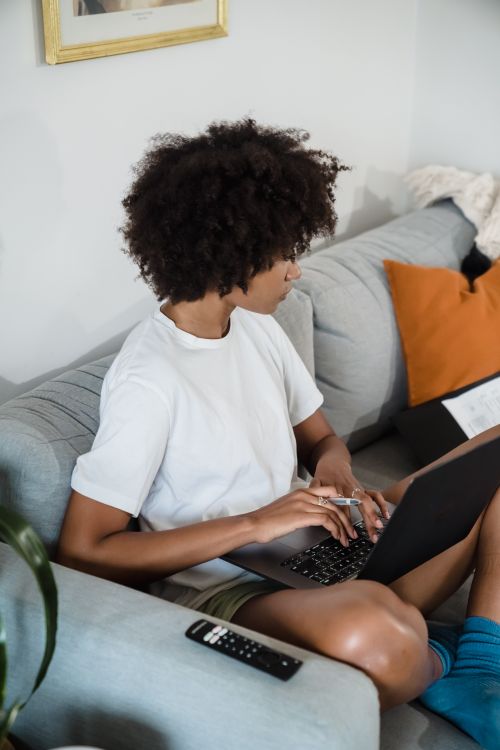 Free Young woman sitting on couch using laptop, showcasing technology and comfort in a home interior. Stock Photo