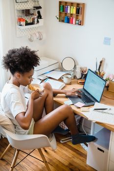 Young woman multitasking at home, eating breakfast while working on a laptop. Cozy and productive home workspace.