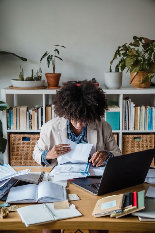 Free Focused woman organizing papers at an office desk with bookshelves and a laptop. Stock Photo