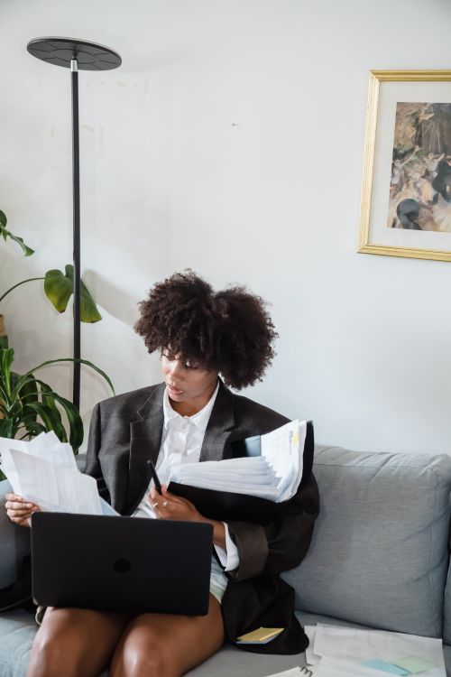 Free Focused woman in home office reviewing papers with laptop on sofa, multitasking. Stock Photo