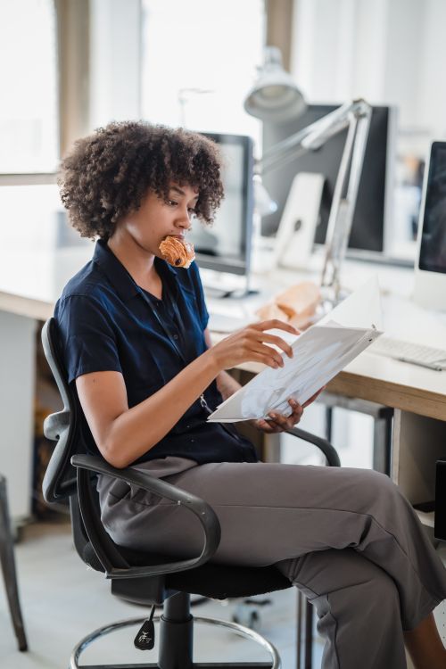 Free A young professional reads documents with a snack in her mouth in a modern office setting. Stock Photo