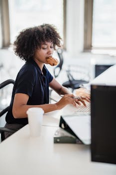 Woman working at desk, eating a croissant while typing on a laptop in a bright office.