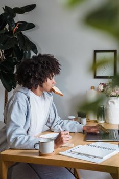 Woman working remotely at desk, enjoying breakfast with toast in mouth.