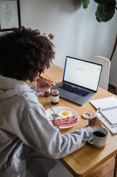 A woman enjoys breakfast at a home office desk with a laptop and coffee.
