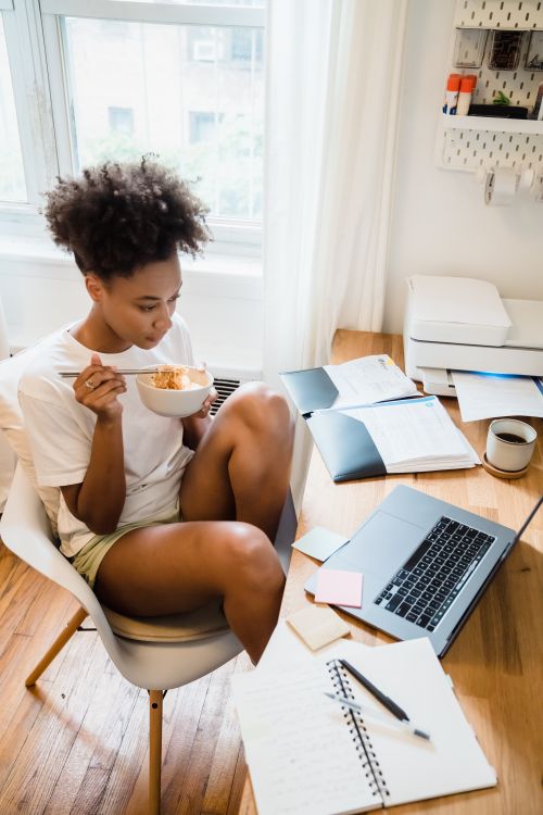 Free Young woman multitasking with breakfast and remote work in a cozy home office setting. Stock Photo