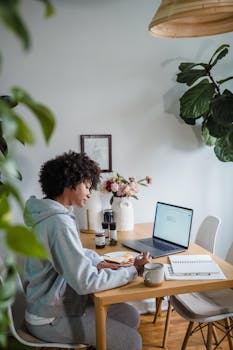 A woman working from home at a table with a laptop, notepad, and coffee, surrounded by plants.