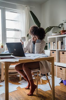 Woman experiences burnout while working remotely, surrounded by papers and a laptop.