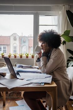 A woman sits at a cluttered desk drinking coffee while working from home.