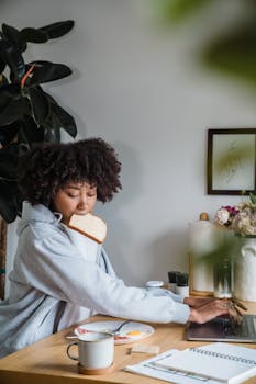 A woman multitasks by eating breakfast and using her laptop in a cozy home office setting.