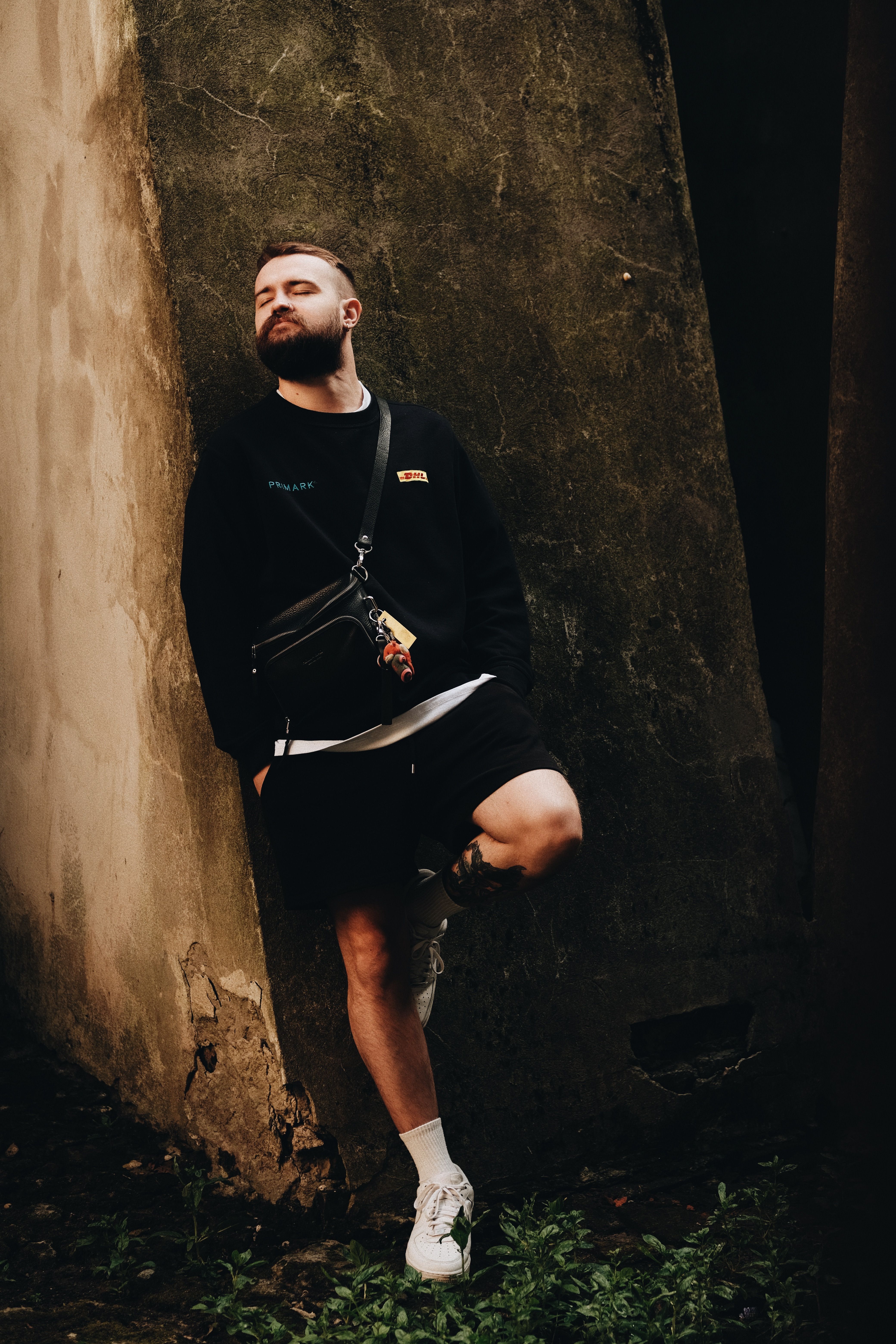 Casual portrait of a young man in a black outfit posing against a textured wall outdoors.
