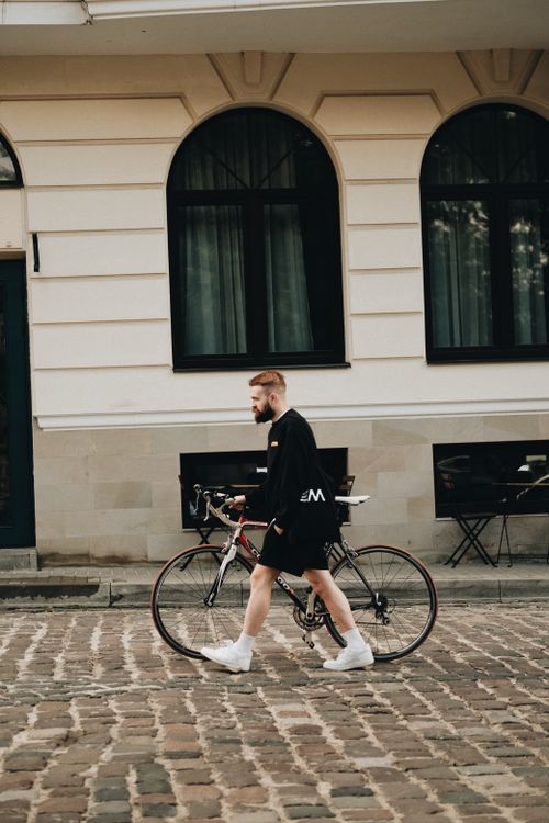 Free A man in urban attire walks a bicycle by a stylish city building. Stock Photo