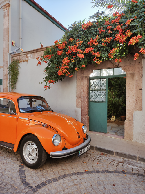 Free Bright orange vintage car parked by a flowering archway in Ayvalık, Turkey. Stock Photo
