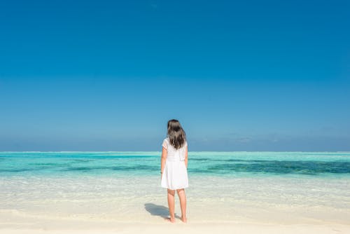 Back View of a Girl in white Dress Standing on the Beach under the Blue Sky