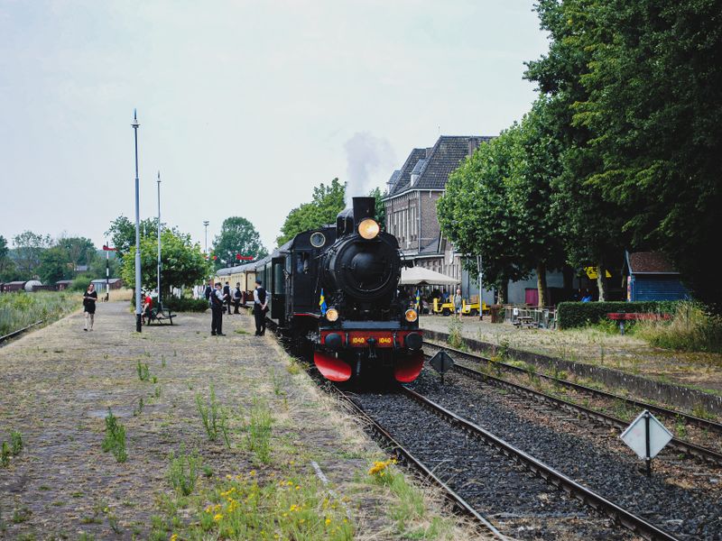 A classic steam locomotive arrives at a rural train station with people and lush greenery around - historic train depots