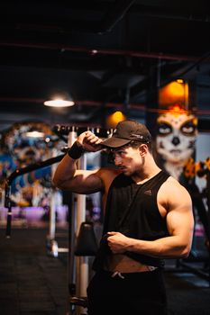 A muscular man in a gym, wearing a black cap and tank top, posing confidently.