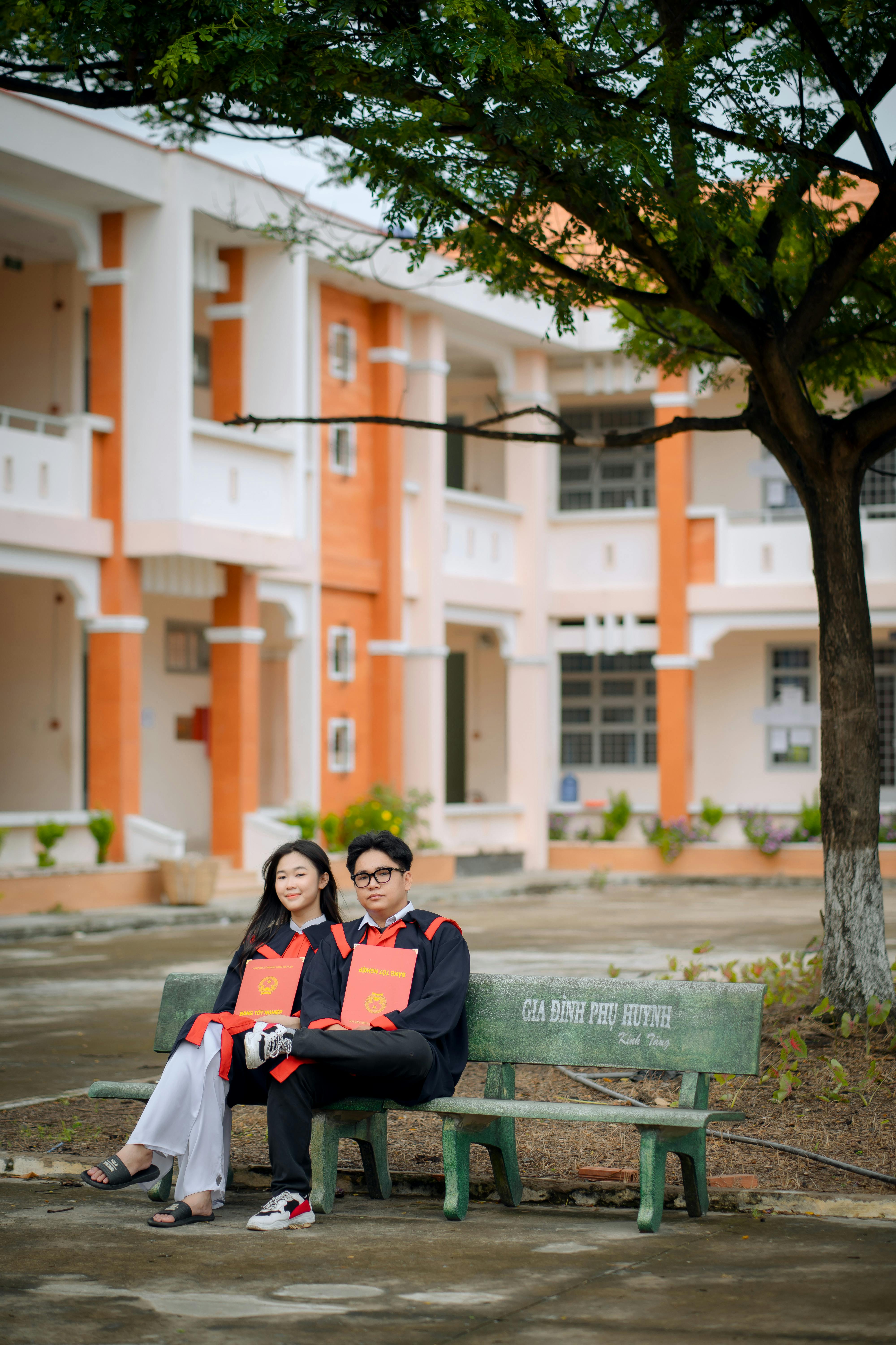 Happy graduates in gowns holding diplomas on campus bench, celebrating their achievement.