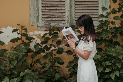 Free Woman in white dress reading a book outdoors near vintage window shutters and foliage. Stock Photo