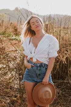 Woman in a field wearing denim shorts and a white shirt, holding a hat.