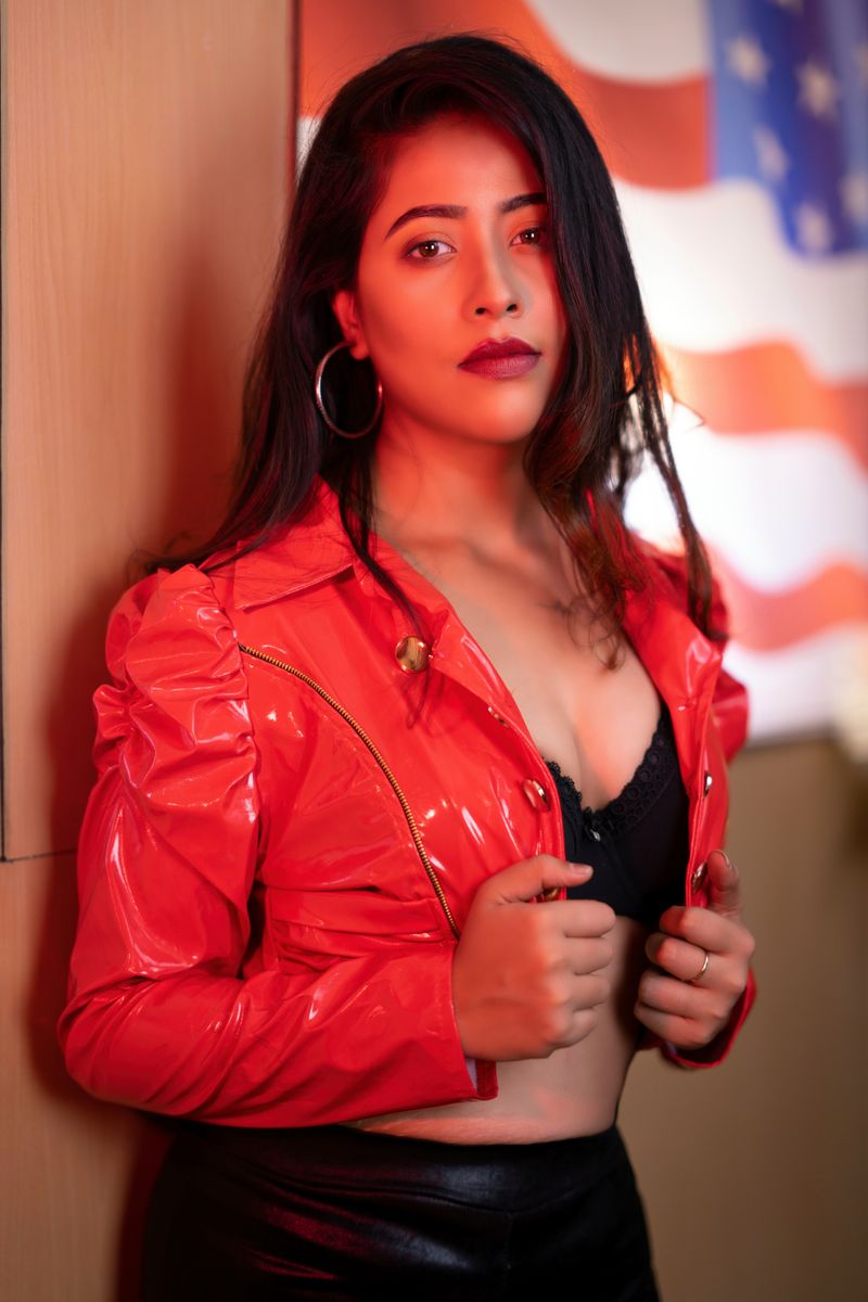 Confident woman in a red leather jacket posing indoors against a blurred flag backdrop.