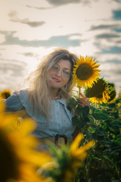 Free Blonde woman in blue dress and glasses poses in a vibrant sunflower field during daytime. Stock Photo