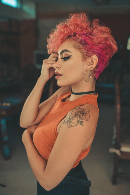 Free Young woman with pink curly hair and floral tattoo, dressed in orange and black, striking a relaxed pose indoors. Stock Photo