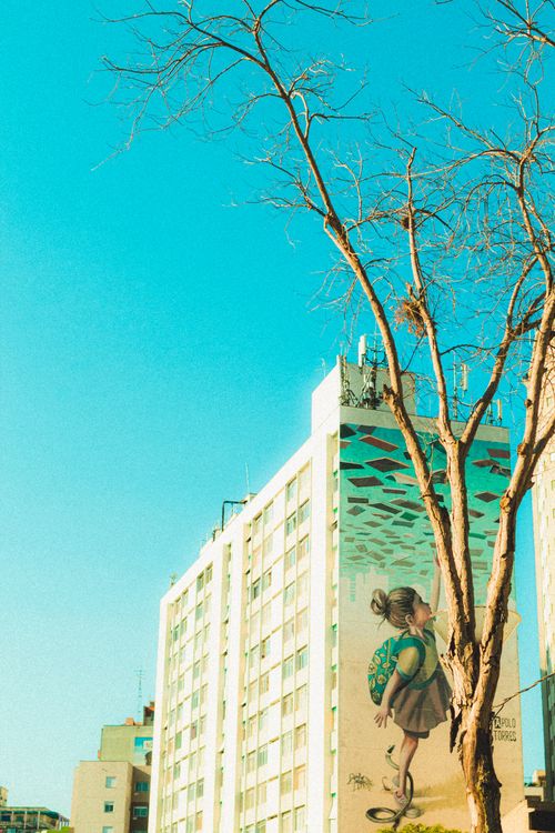 Free A vibrant urban scene featuring a bare tree against a building with a large mural, under a clear blue sky. Stock Photo