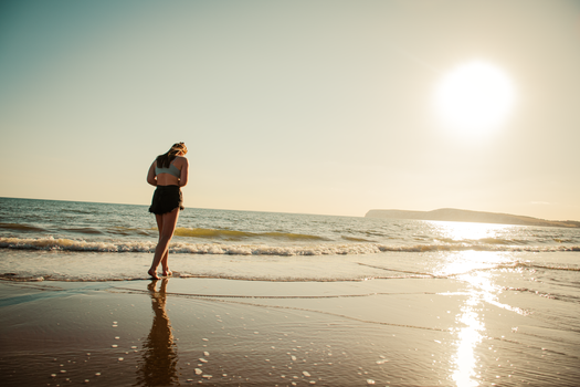A woman walks along a sunlit beach in England during a serene sunset.