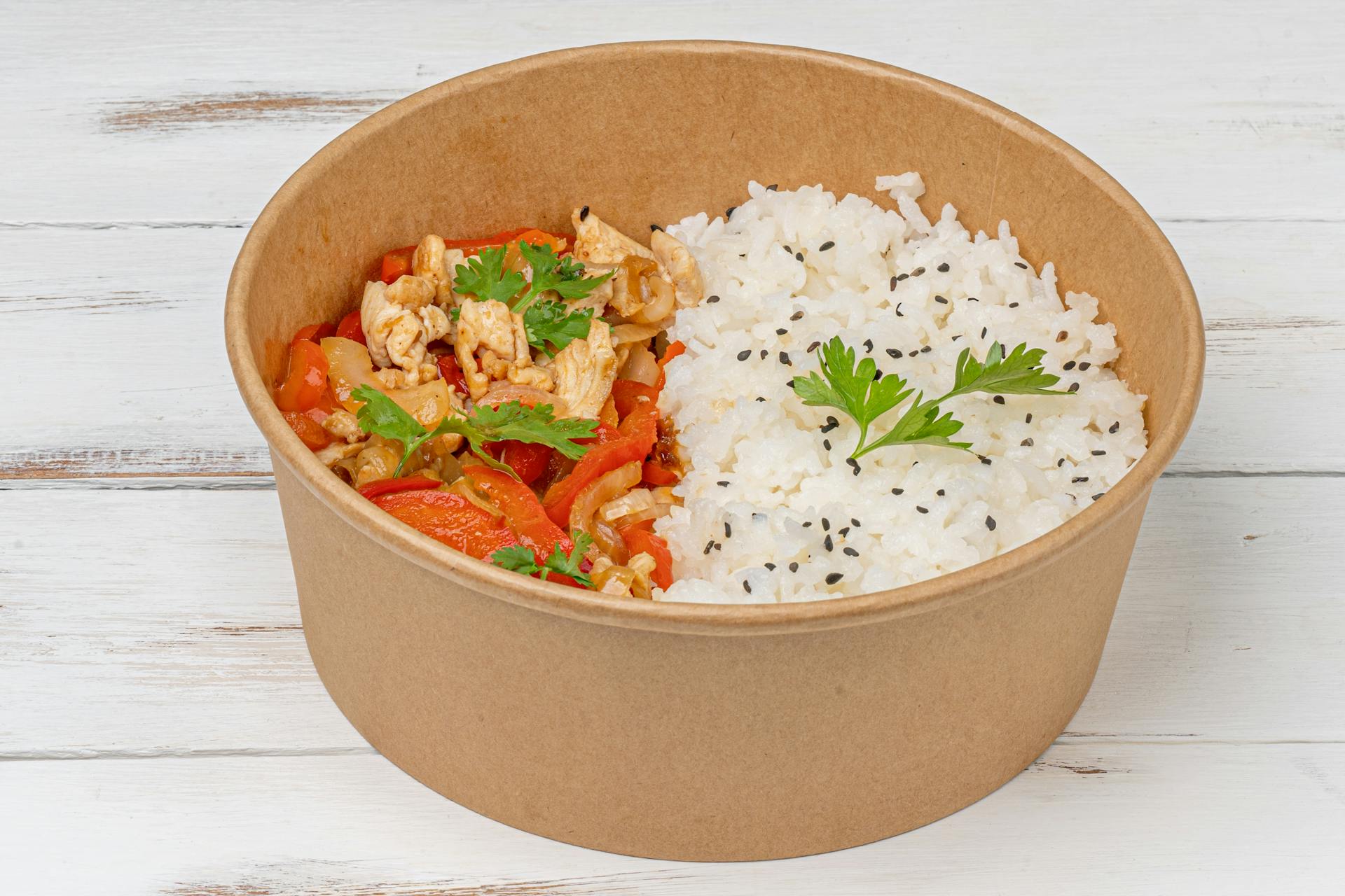 A delicious eco-friendly takeout rice bowl with vegetables in a paper container on a wooden table.
