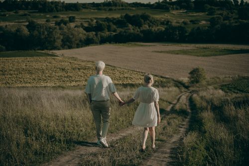 Free An elderly couple enjoys a peaceful walk through lush countryside fields, holding hands under the warm summer sun. Stock Photo