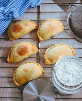 Freshly baked Peruvian empanadas on a cooling rack with a jar of flour nearby.