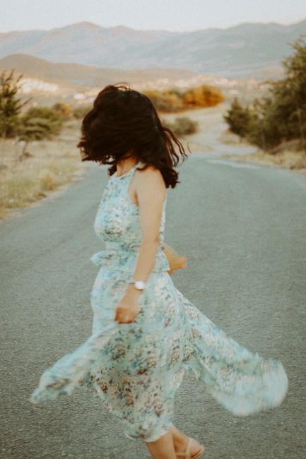 A woman in a blue floral dress twirls on an open road surrounded by scenic hills.