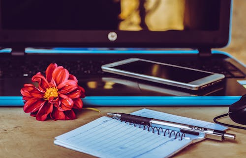 White Smartphone on Black Laptop Next to Pen on White Notepad